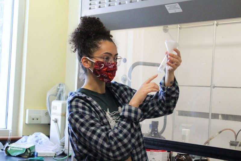 A young woman wearing a face mask in a lab holds up, looks at, and points to a test tube.